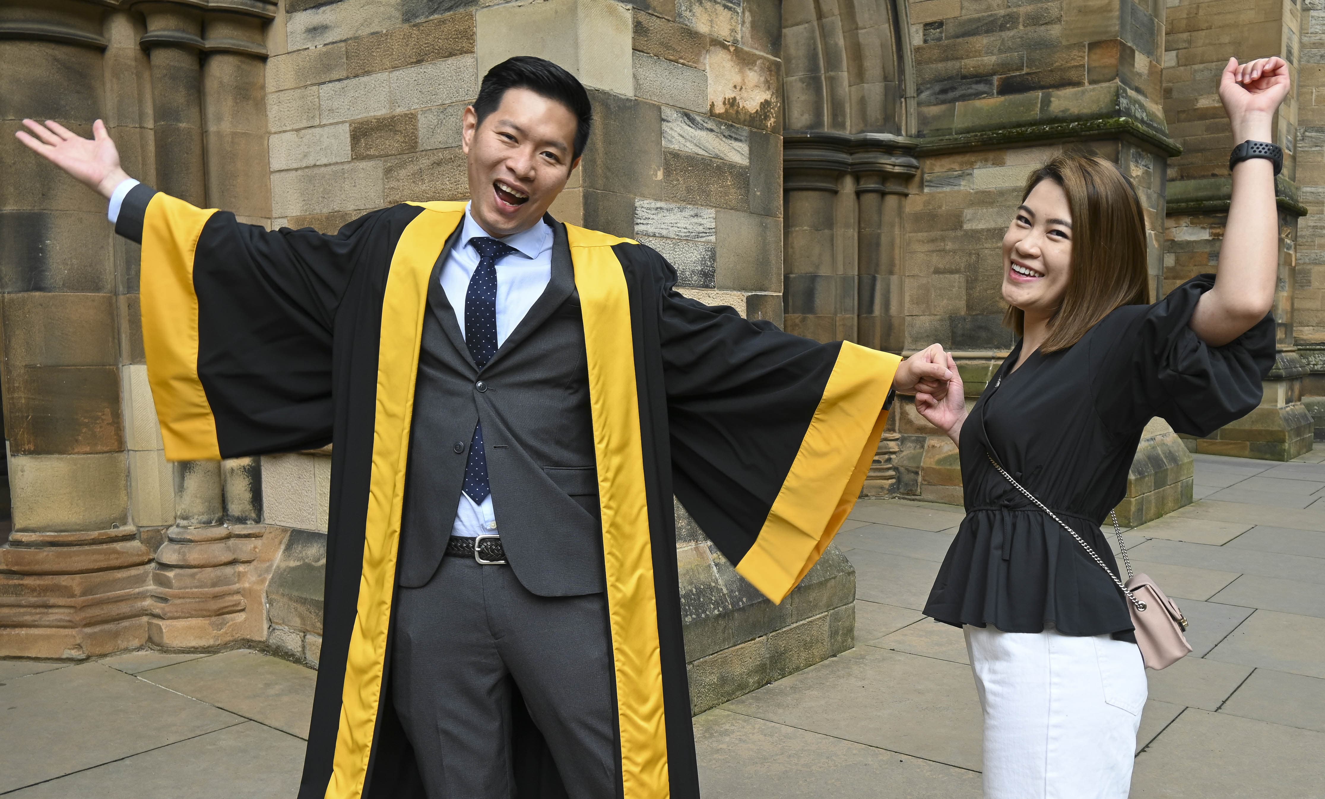 A smiling graduate in ceremonial academic robes stands with arms outstretched next to a woman celebrating with raised arms outside the Royal College of Physicians and Surgeons of Glasgow.