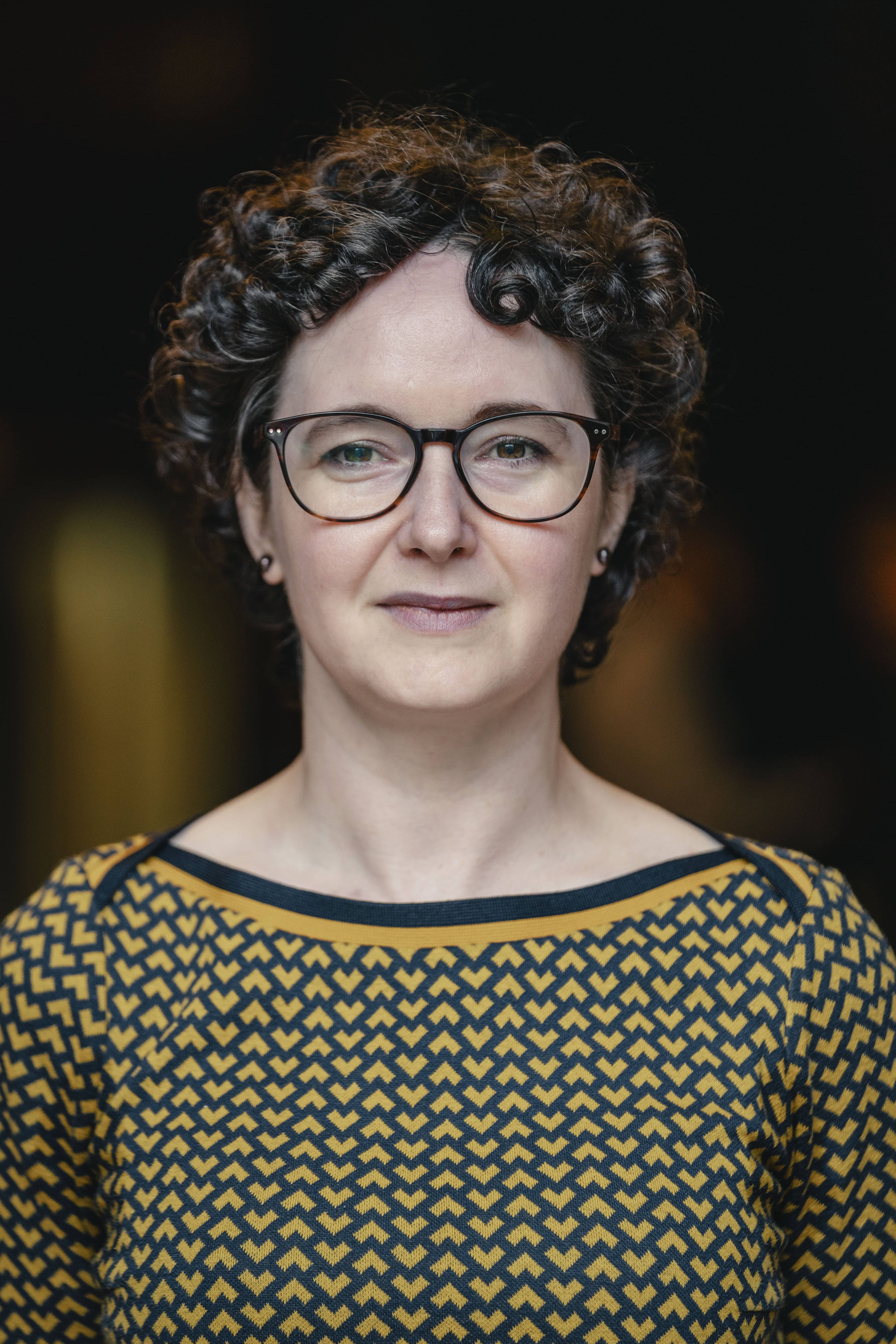 Portrait of a woman with short curly dark hair and glasses, wearing a patterned mustard and navy top, standing against a dark blurred background.