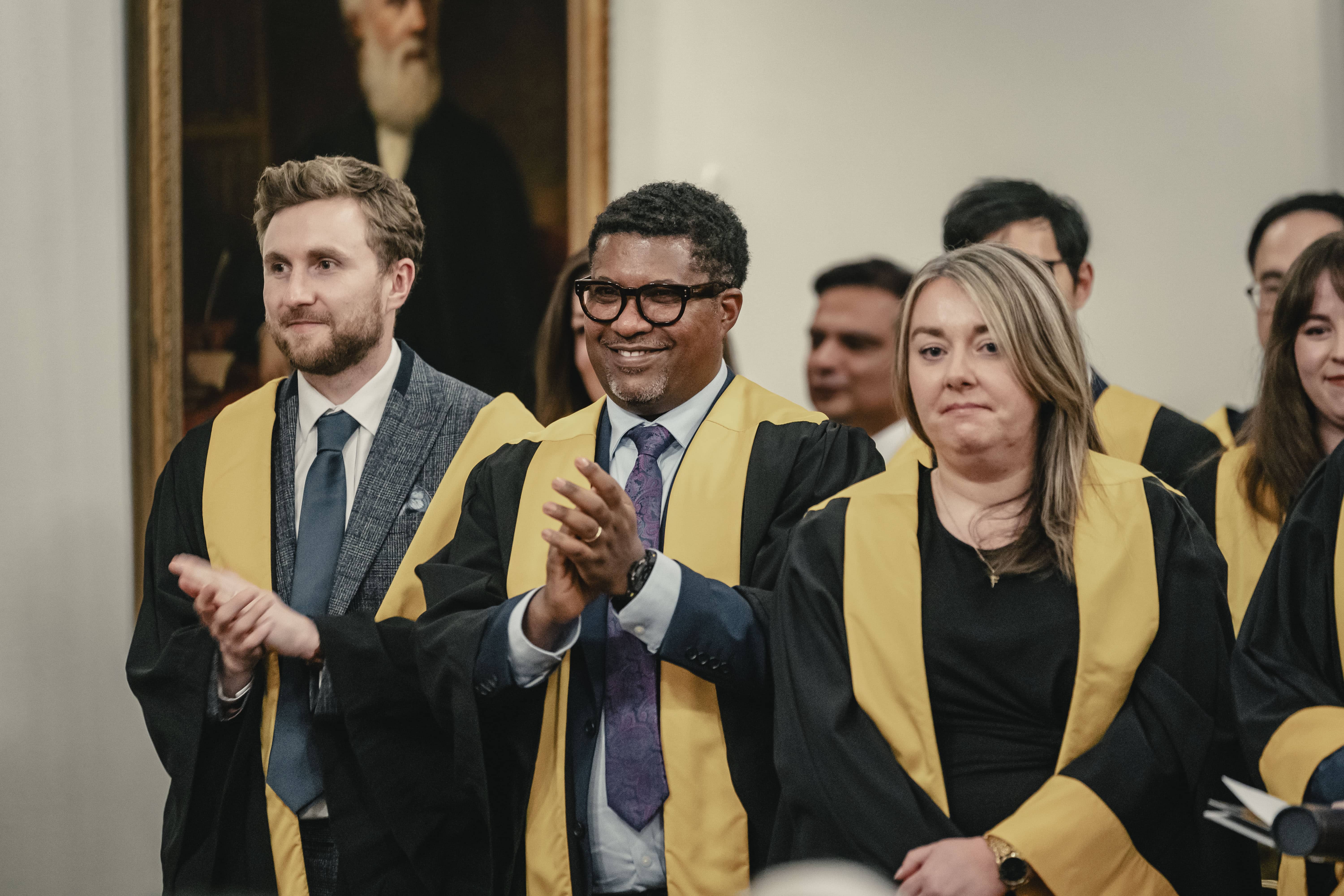 Group of graduates in ceremonial gowns applauding during a formal indoor event, with a portrait painting in the background.